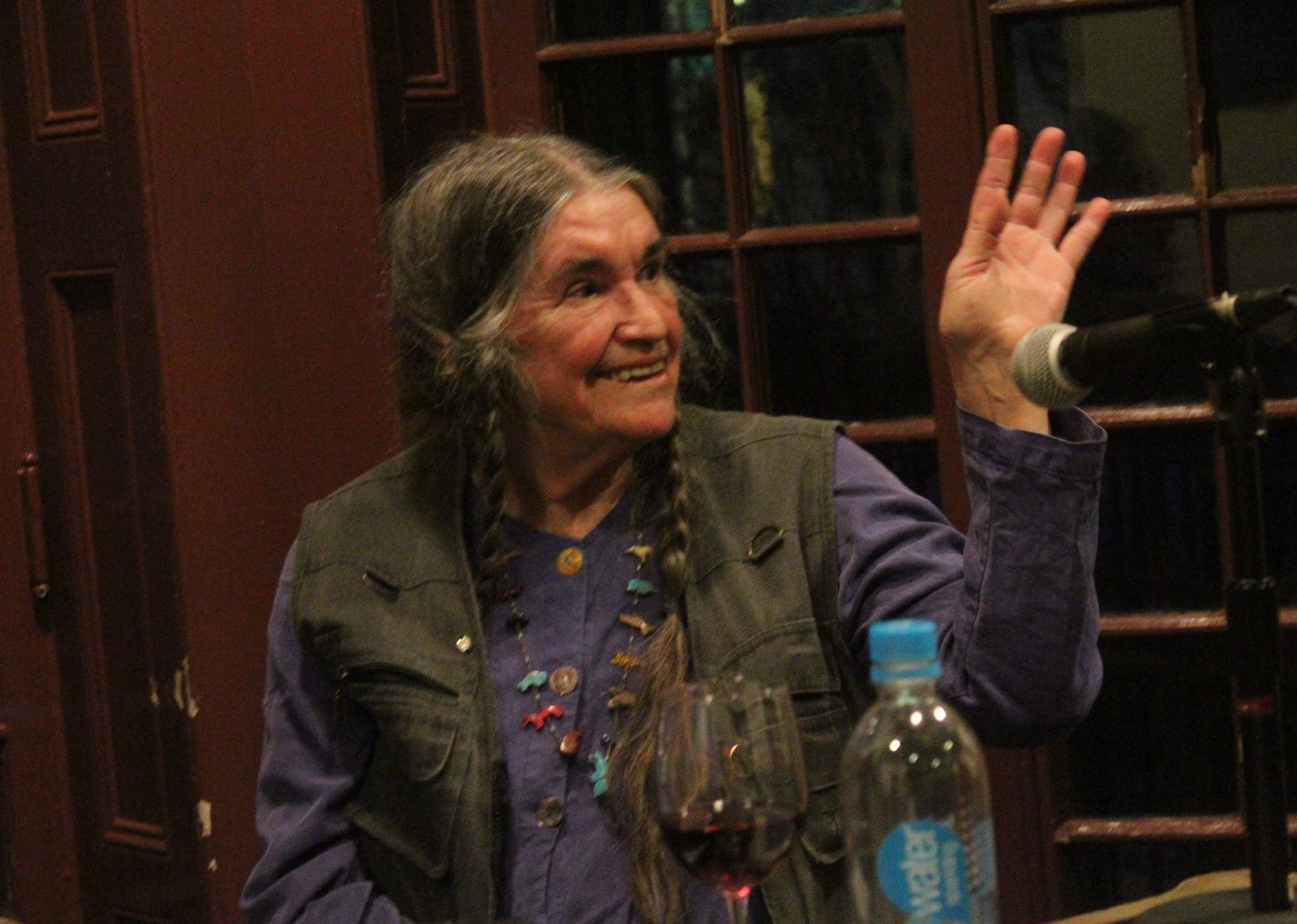 Bernadette Mayer sitting and waving before a microphone with a glass of wine and a water bottle.